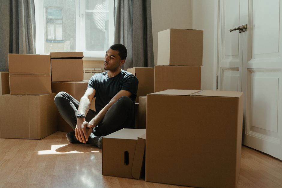 Young child sits thoughtfully on the floor of a home, surrounded by packed cardboard moving boxes, representing the emotio...